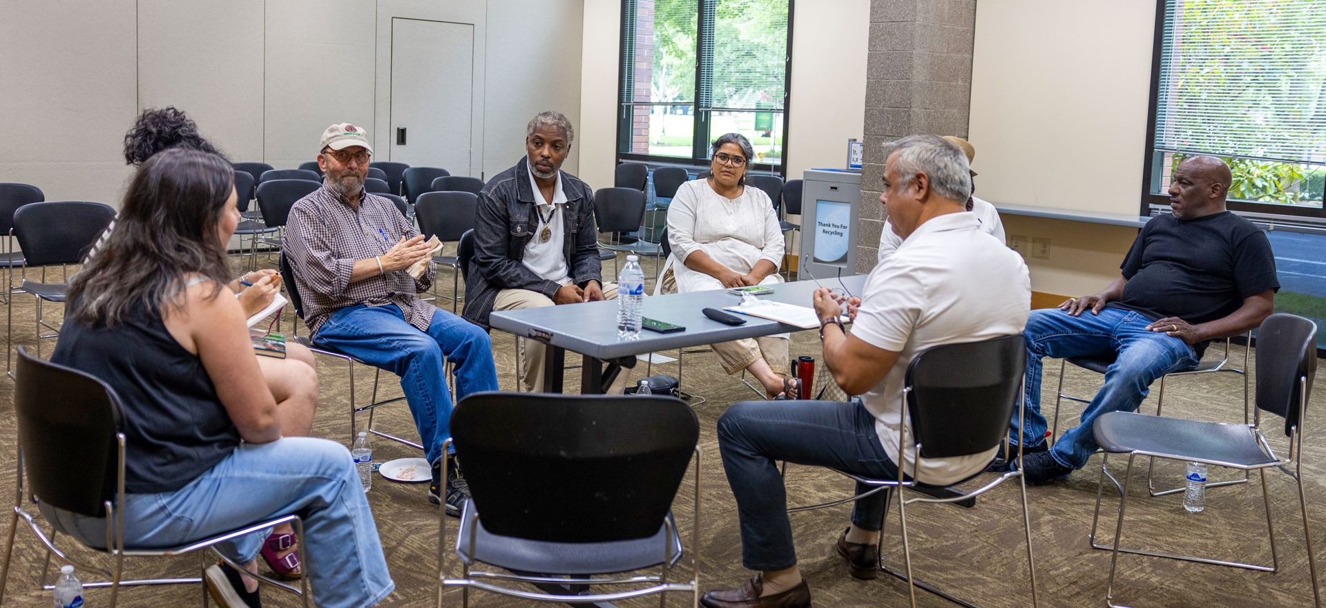 A Lara Media Services facilitator in a white shirt engages in active dialogue with a diverse group of Beaverton community members seated in a circle during a multicultural gap analysis session. Another Lara Media team member takes notes on the left.